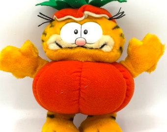 Könnte beinhalten: Orangefarbenes Plüschtier von Garfield der Katze, verkleidet als Kürbis. Das Spielzeug hat einen grünen, blattreichen Stiel auf dem Kopf und ein breites Grinsen.