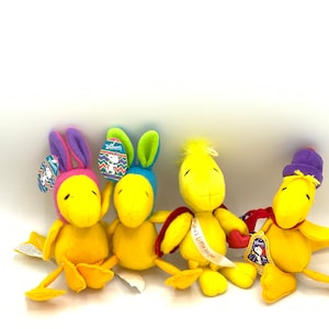 May include: Four plush yellow chicks with different colored hats and accessories. The chicks are wearing bunny ears, a purple hat, a red and white sash, and a blue hat with an Easter egg design. The chicks are all sitting on a white surface.