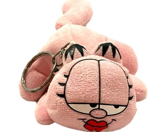 Può includere: Un portachiavi Garfield in peluche rosa. Il portachiavi presenta il volto di Garfield con dettagli neri, occhi chiusi e labbra rosse. Il portachiavi ha un anello di metallo argentato attaccato al peluche.