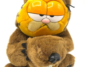 Può includere: Un peluche di Garfield il gatto vestito da orso bruno. Garfield indossa un costume da orso bruno con un cappotto di pelliccia marrone, zampe marroni e una testa di orso bruno. Il viso di Garfield è visibile attraverso il costume da orso.
