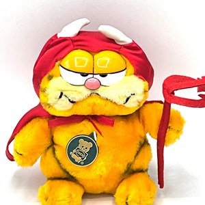 May include: A plush orange Garfield cat dressed as a devil for Halloween. He is wearing a red cape and horns, and holding a red pitchfork. The plush toy has a Dakin tag attached to it.