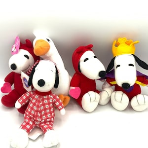 May include: A collection of Snoopy plush toys in various outfits, including a red devil costume, pajamas, and a king's attire. A white goose plush is also present. The toys are on a white background.