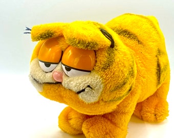 May include: A plush Garfield toy with a yellow and orange striped pattern. The toy has a grumpy expression with large, round eyes and a pink nose.