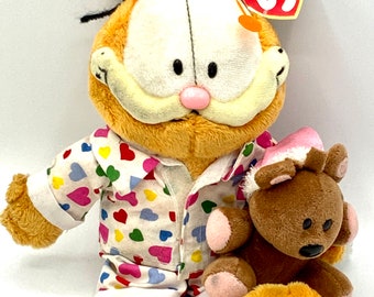 Könnte beinhalten: Ein plüschiges orangefarbenes Garfield-Spielzeug, das einen weißen und rosafarbenen Schlafanzug mit bunten Herzen trägt. Es hält einen braunen Teddybären in seinen Armen. Das Spielzeug hat ein Ty Beanie Babies-Etikett an seinem Ohr.