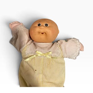 May include: A doll with a round, tan vinyl head, dark eyes, and a small mouth. The doll wears a white top with lace trim and a yellow and white pinstriped bottom, featuring a small yellow ribbon bow at the center of the chest area.