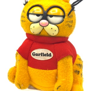 Peut inclure: Un jouet en peluche Garfield portant un pull rouge avec le mot "Garfield" dessus. Le jouet a des yeux noirs, un nez rose et un corps rayé jaune et orange.