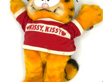 May include: A plush Garfield toy wearing a red shirt with the text "Kissy, Kissy" in white with a red heart on the shirt. The toy has orange fur, black stripes, and a black nose.