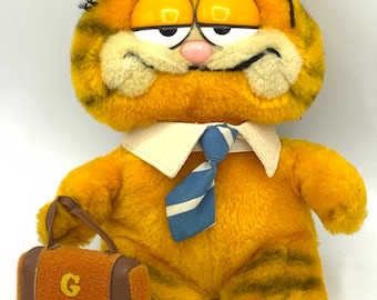 May include: A plush orange and black Garfield cat wearing a white shirt and blue and white striped tie. The cat is holding a brown suitcase with a capital G on the front.