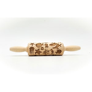 May include: A wooden rolling pin with a sea life design. The rolling pin is engraved with images of a starfish, a pufferfish, a jellyfish, a turtle, a seahorse, seaweed, and fish. The rolling pin is light brown and has a natural wood finish.