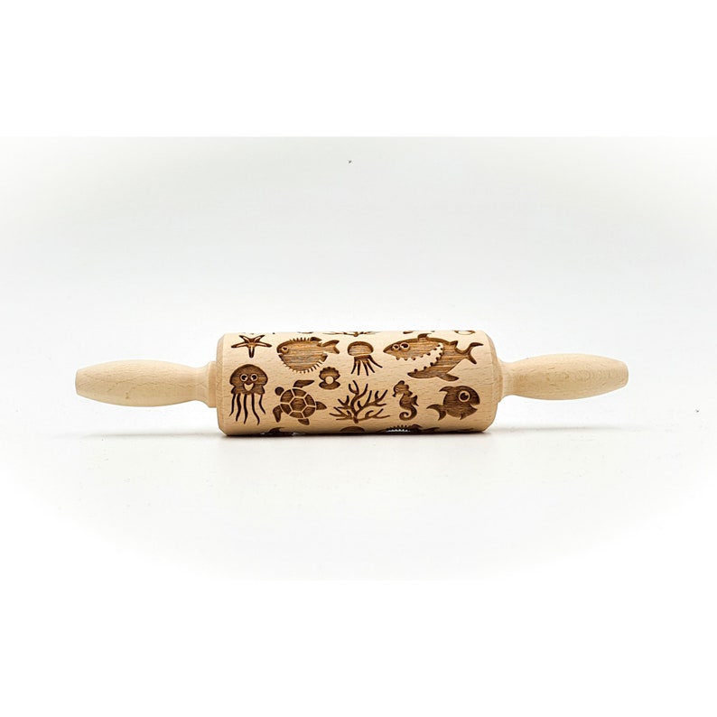 May include: A wooden rolling pin with a sea life design. The rolling pin is engraved with images of a starfish, a pufferfish, a jellyfish, a turtle, a seahorse, seaweed, and fish. The rolling pin is light brown and has a natural wood finish.