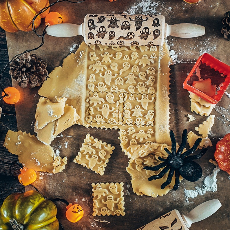 Peut inclure: Un gros plan d'un rouleau &agrave; p&acirc;tisserie avec un motif de fant&ocirc;me, utilis&eacute; pour cr&eacute;er des biscuits sur le th&egrave;me d'Halloween. Le rouleau &agrave; p&acirc;tisserie est sur une surface brune avec de la p&acirc;te &agrave; biscuits, une araign&eacute;e et de petites citrouilles.