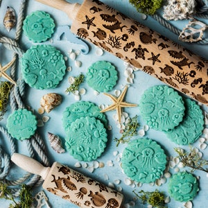 May include: Two wooden rolling pins with engraved designs of sea creatures and plants. The rolling pins are on a blue background with green moss, white seashells, and blue and green cookie dough cutouts in the shapes of sea creatures and plants.