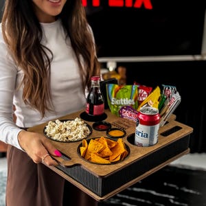Può includere: Un vassoio in legno con scomparti per popcorn, patatine e salse, insieme a una bottiglia di soda e una lattina di Aria. Il vassoio contiene anche varie caramelle e patatine in formato snack. Il vassoio è tenuto da una persona.