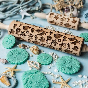 May include: Two wooden rolling pins with an ocean theme design. The rolling pins are engraved with images of sea creatures, such as jellyfish, seahorses, and fish. The rolling pins are surrounded by blue and white cookies with the same ocean theme design. The cookies are on a light blue surface with white seashells and a starfish.