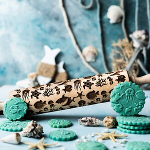 May include: A wooden rolling pin with a sea life design. The rolling pin is engraved with images of fish, seahorses, starfish, seashells, and coral. There are several green sugar cookies with the same sea life design on a light blue surface.