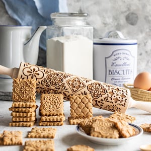 Op de afbeelding: Een houten deegroller met een reliëf bloemmotief, naast stapels ingewikkeld ontworpen koekjes. Een witte kan, bloem en een koektrommel staan op de achtergrond. De koekjes zijn lichtbruin en hebben gedetailleerde patronen.