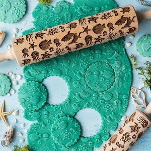 May include: A wooden rolling pin with a carved design of sea creatures and coral. The rolling pin is used to create impressions in green dough. The dough is cut into circles and has the same sea creature and coral design. The rolling pin is on a light blue surface with white seashells and a starfish.