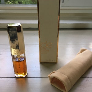 May include: A gold and white perfume bottle with a gold cap and a small, gold-colored atomizer. The bottle is labeled "Cie Pure Perfume Cologne 1/3 FL OZ". The bottle is sitting on a white surface next to a small, yellow fabric pouch.