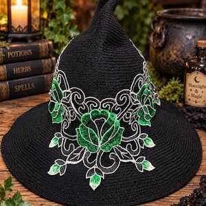 May include: A black pointed hat featuring intricate white and green floral embroidery. The design showcases detailed green rose motifs surrounded by white swirling patterns that wrap around the base of the crown, set against a solid black fabric.