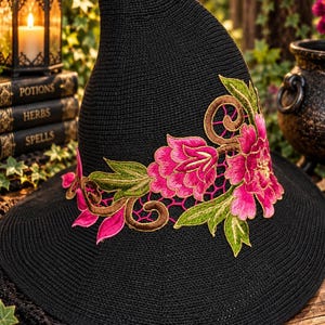 Puede incluir: Un sombrero negro de punta con ala ancha, que presenta un intrincado bordado de flores de color rosa vibrante, hojas verdes y enredaderas marrones. El sombrero está construido con un material tejido texturizado con una copa alta y ligeramente curva.