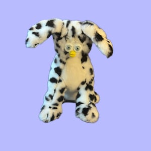 Puede incluir: Un juguete de peluche negro y blanco con manchas que se asemeja a un perro con ojos grandes y redondos y un pico amarillo. El juguete está sentado sobre un fondo morado claro.