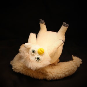 Puede incluir: Un peluche beige claro con una textura difusa y ojos grandes y expresivos. El juguete tiene un botón amarillo y detalles negros en los pies. El juguete está tumbado boca arriba sobre un fondo negro.