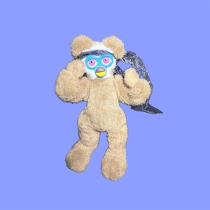 Puede incluir: Un juguete de peluche marrón con una cara tipo Furby, que lleva una diadema negra y blanca y una capa negra. El juguete tiene ojos rosas y azules y un pico amarillo. El fondo es de color púrpura claro sólido.