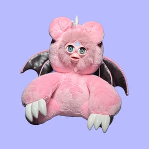 Puede incluir: Un peluche rosa con orejas grandes, ojos azules y garras blancas. El juguete tiene alas negras y grises y un pequeño cuerno. El juguete es un peluche único y personalizado.