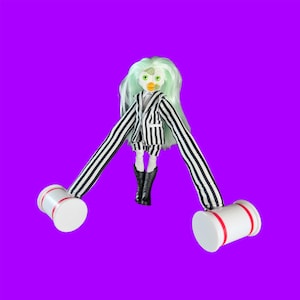 Puede incluir: Una muñeca Furby personalizada con cabello verde, vestida con un traje a rayas blanco y negro y botas negras. La muñeca tiene brazos largos que terminan en mazos blancos con rayas rojas, sobre un fondo púrpura.