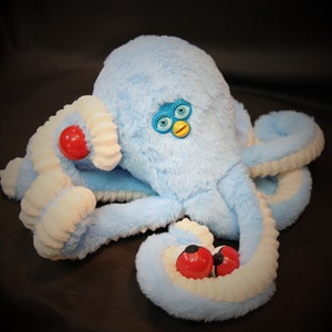 Puede incluir: Un juguete de peluche de pulpo azul claro con tentáculos blancos y azul claro. El juguete tiene ojos redondos rojos y un pequeño detalle de cara azul y amarilla. El peluche es suave y esponjoso.