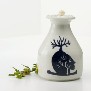May include: A white ceramic oil lamp with a blue and black design of a tree growing out of a person's head. The lamp has a wick and a white cap.