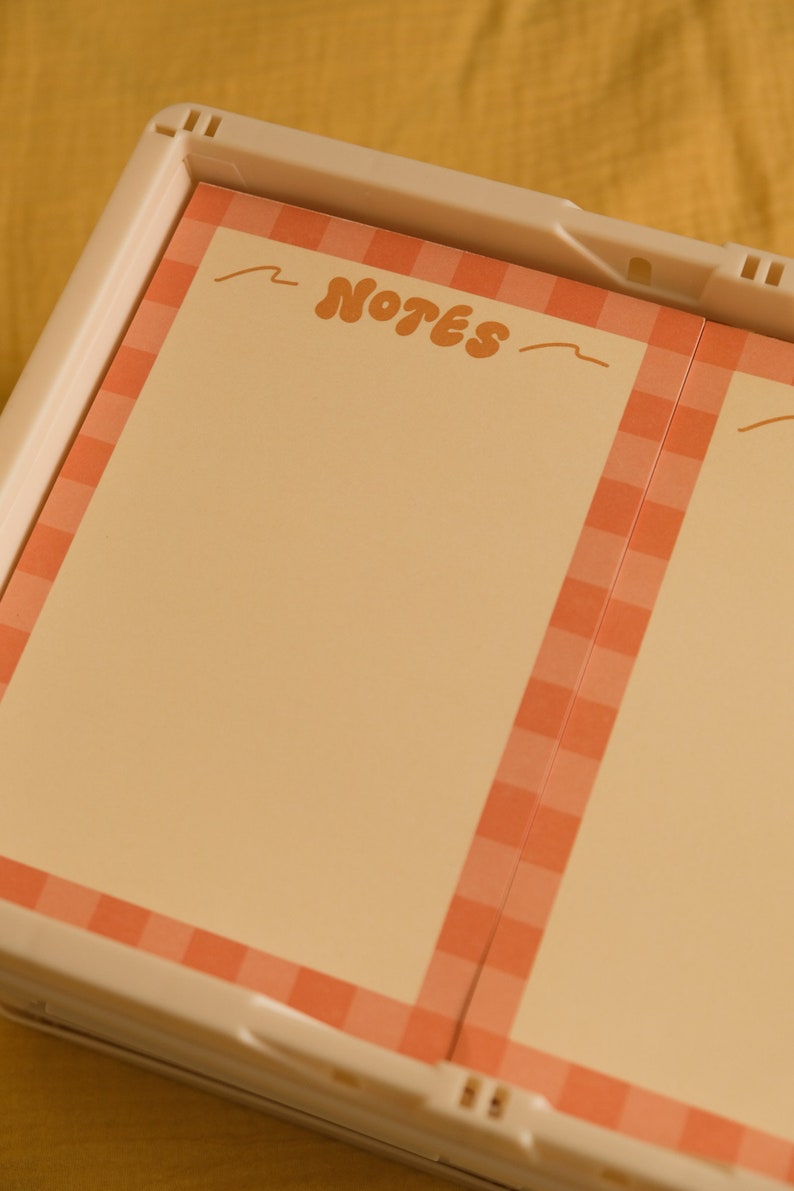 Może przedstawiać: Notatnik z jasnym, brzoskwiniowym tłem i brzoskwiniowo-białą obw&oacute;dką w kratkę vichy. Słowo "NOTES" jest napisane w stylu retro. Notatnik znajduje się w białej plastikowej ramie.