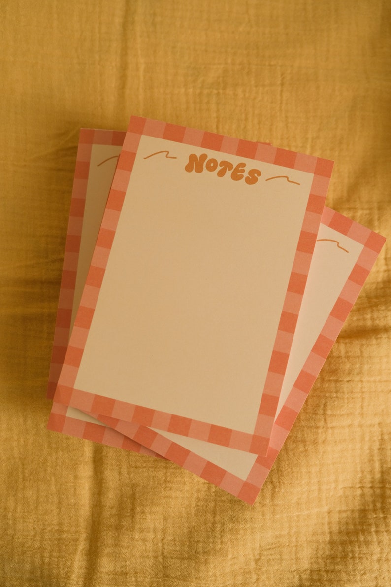 Może przedstawiać: Trzy ułożone w stos notatniki z brzoskwiniowym tłem i brzoskwiniowo-białą obw&oacute;dką w kratę. Słowo "NOTES" jest napisane w stylu retro na g&oacute;rze notatnika. Notatniki leżą na ż&oacute;łtej, teksturowanej powierzchni.