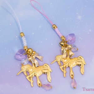 May include: Two gold-colored unicorn charms with pink and white cords. Each charm features a unicorn design with a bow, a bell, and a dangling crystal. The background is a soft gradient of purple and blue.
