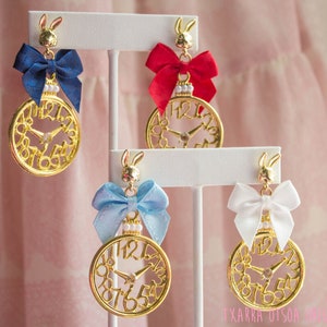 May include: Gold-colored earrings with a clock face design, featuring a rabbit charm and a satin bow in blue, red, light blue, and white. The earrings have a pearl accent and are displayed on white stands.