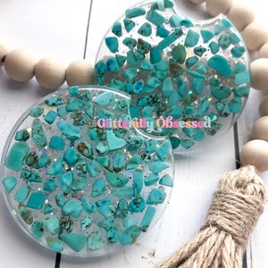 May include: Two turquoise blue and white car coasters with a glittery finish. The coasters are round and have a variety of turquoise blue chips embedded in the resin. The coasters are sitting on a white wooden surface with a brown tassel.