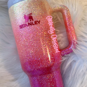 May include: A glittery Stanley tumbler in a gradient of pink and orange hues. The tumbler features a handle and a pink lid. The Stanley logo is visible, along with the text "Glitterally Obsessed".