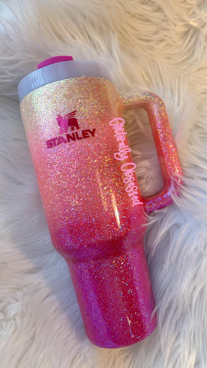 May include: A glittery Stanley tumbler in a gradient of pink and orange hues. The tumbler features a handle and a pink lid. The Stanley logo is visible, along with the text "Glitterally Obsessed".