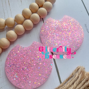 May include: Two pink glitter-covered round coasters with a slightly rounded edge. The coasters are on a white wooden surface. The text "Literally obsessed" is written in pink, blue, and yellow letters on the surface.