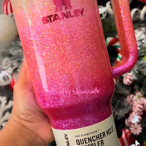 May include: A glittery Stanley Quencher H2.0 tumbler in a gradient of pink and gold. The 40 oz (1.18 L) tumbler has a handle and a lid. The text "STANLEY" and "glitterallyobsessed" are visible.