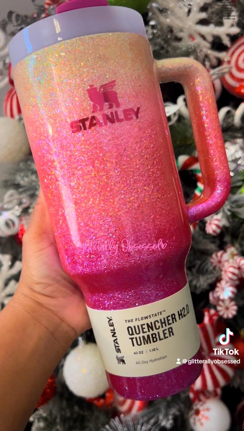 May include: A glittery Stanley Quencher H2.0 tumbler in a gradient of pink and gold. The 40 oz (1.18 L) tumbler has a handle and a lid. The text "STANLEY" and "glitterallyobsessed" are visible.