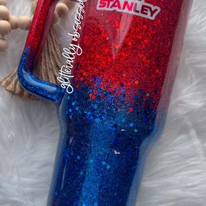 May include: A glittery tumbler in red and blue with a handle. The top is red, transitioning to blue at the bottom. A white and pink Stanley logo is visible. The tumbler is designed for cold beverages.