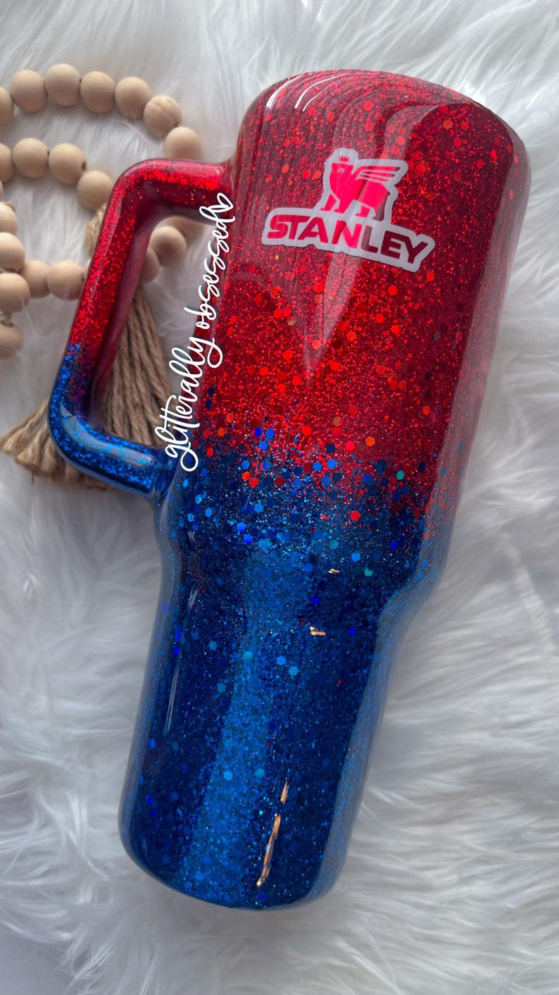 May include: A glittery tumbler in red and blue with a handle. The top is red, transitioning to blue at the bottom. A white and pink Stanley logo is visible. The tumbler is designed for cold beverages.