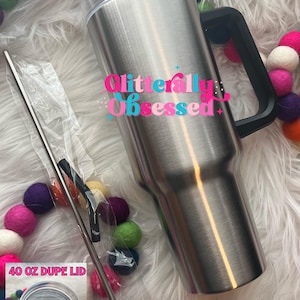 May include: A stainless steel 40 oz tumbler with a black handle and lid, accompanied by a metal straw and a lid. The text "40 OZ DUPE" and "Glitterally Obsessed" are visible. Colorful felt ball garland decorates the scene.