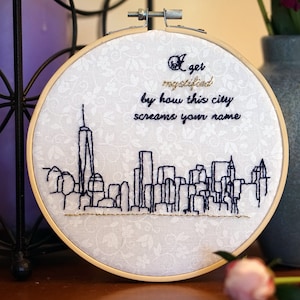 May include: Embroidered hoop art depicting the New York City skyline in dark blue thread. The text "I get mystified by how this city screams your name" is stitched above the skyline. The fabric has a white floral pattern.
