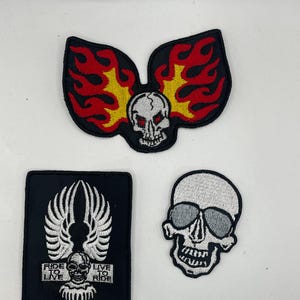 May include: Three embroidered patches featuring skull designs. One patch shows a skull with flames, another has a skull with sunglasses, and the third has a skull with wings and the text "RIDE TO LIVE, LIVE TO RIDE".