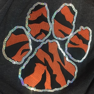 May include: A black fabric with a large orange and black tiger paw print embroidered with silver sequins.
