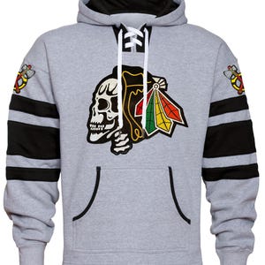 May include: A gray hooded sweatshirt with black and white stripes on the sleeves. The front of the sweatshirt features a black and white skull with a red, green, and yellow logo. The logo is a stylized Native American headdress.