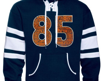 Könnte beinhalten: Ein marineblauer Hoodie mit weißen Streifen an den Ärmeln. Der Hoodie hat eine Kapuze mit Kordelzug und einen Schnürverschluss vorne. Die Nummer 85 ist in Orange auf der Vorderseite des Hoodies aufgedruckt, wobei die Nummer aus Namen berühmter Fußballspieler besteht.