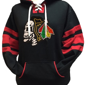 Puede incluir: Sudadera con capucha negra con mangas a rayas rojas y negras. La sudadera tiene un cuello con cordones y un gran diseño bordado de calavera y plumas en la parte delantera.